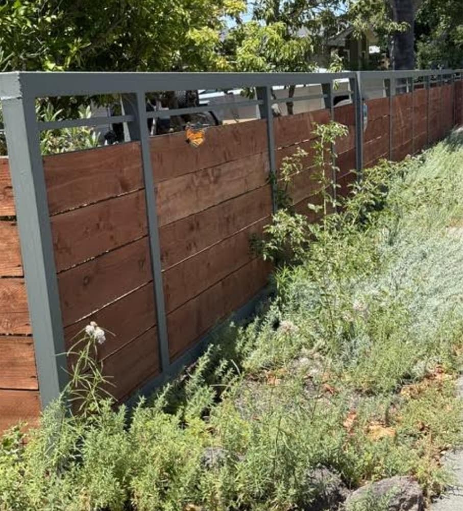 A native-planted garden bed along a residential fence line, with a butterfly resting on the fence rail.